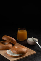 A wooden board of bagels with a glass of orange juice and a butter knife. Scene is casual and relaxed, as it seems like someone is having a simple breakfast