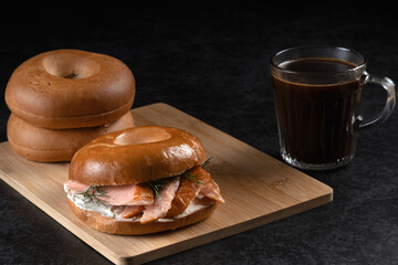 A sandwich with salmon and dill is on a wooden board next to a mug of coffee. The sandwich is placed on top of wooden board and the coffee is in a glass. Concept of comfort and relaxation