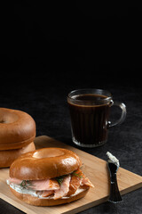 A sandwich and a cup of coffee are on a wooden table. The sandwich is a bagel with salmon and dill. The coffee is black and in a glass. Scene is casual and relaxed