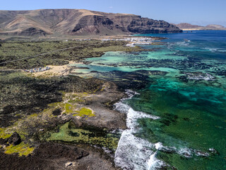 Fototapeta premium Aerial view of volcanic reef patterns and turquoise shallow waters near Órzola, Lanzarote, with mountains and coastline in the background. 