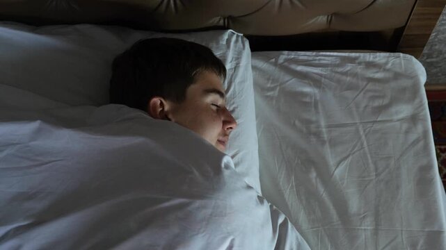 Top view of a teenage boy sleeping on his side on a bed with white bedding. The boy sleeps calmly and peacefully, smiling slightly in his sleep. Comfortable bed and orthopedic mattress.
