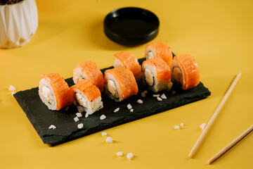 Sushi rolls arranged neatly on a black slate platter with dipping sauce on a yellow background