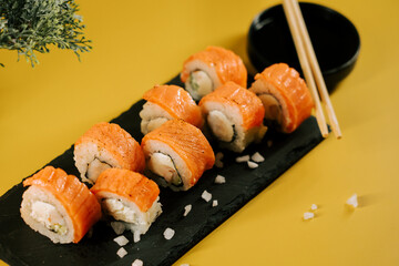 Sushi rolls served on a black stone plate with chopsticks and soy sauce on a yellow background