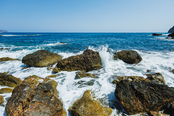 Waves crash over coastal rocks at midday with bright sunlight illuminating the turquoise sea and clear skies in a tranquil natural landscape