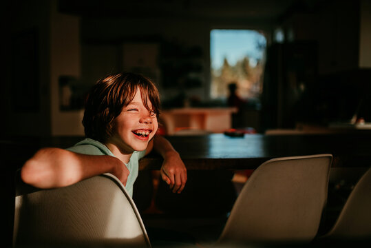 Boy sitting at a dining room table laughing with sunlight on his face