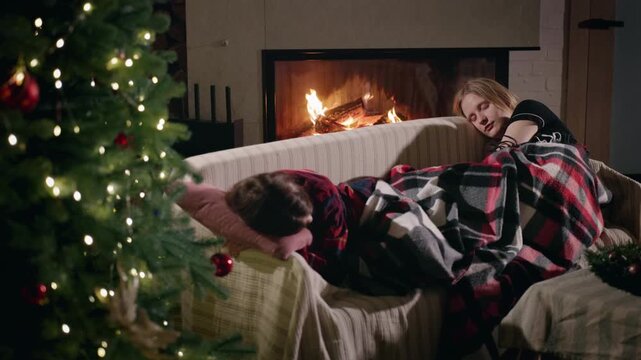 Two teenage girls fall asleep together on a cozy sofa under a checkered blanket, with a Christmas tree glowing softly beside them and a warm fire burning in the fireplace.