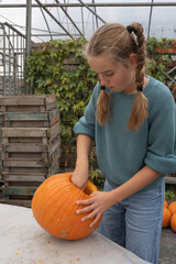 Young girl with braided hair, carving an orange pumpkin in a greenhouse, surrounded by wooden crates and vibrant green plants, showcasing autumn creativity and seasonal activities