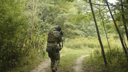 Camouflaged and masked army soldier with AR15 carbine on a mission. Carefully sneaking through a thick green forest then pausing to take aim. Military recon