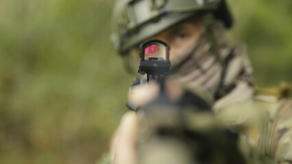 Camouflaged and masked army soldier with AR15 carbine on a mission. Rifle with a collimator aiming at the camera. Selective focus. Military recon