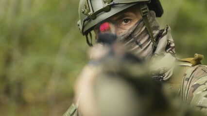 Camouflaged and masked army soldier with AR15 carbine on a mission. Rifle with a red collimator aiming at the camera. Selective focus. Military recon