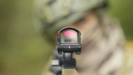 Camouflaged and masked army soldier with AR15 carbine. Rifle with a red collimator aiming at the camera. Selective focus. Military recon