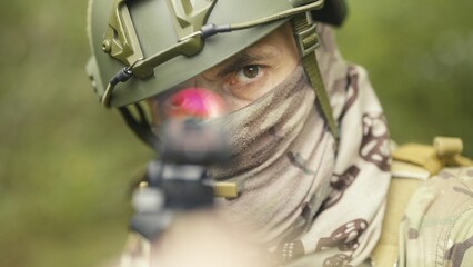Camouflaged and masked army soldier with AR15 carbine. Rifle with a collimator aiming at the camera. Selective focus. Military recon