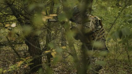 Camouflaged army soldier with AR15 carbine on a mission. Traversing the rough terrain of trees and bushes and taking aim. Military recon
