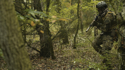 Camouflaged army soldier with AR15 carbine on a mission. Traversing the rough terrain of trees and taking aim. Military recon