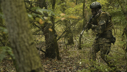 Camouflaged army soldier with AR15 carbine on a mission. Traversing the rough terrain of trees and taking aim. Military recon