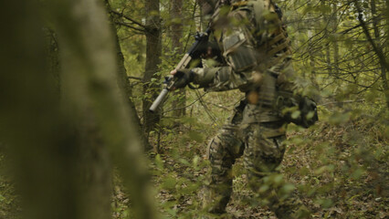Camouflaged army soldier with AR15 carbine on a mission. Traversing the rough terrain of lush trees and bushes. Military recon