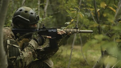 Camouflaged army soldier with AR15 carbine on a war recon mission. Hiding in green bushes and leaves to aim at target. Military recon