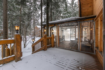 A charming cabin sits amid snow-covered trees, featuring a wooden deck with warm lanterns. The serene setting captures a quiet winter day, perfect for relaxation and enjoying nature.
