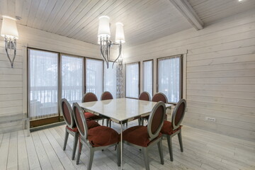 A spacious dining area features a large rectangular table surrounded by red upholstered chairs. Large windows allow natural light to fill the wooden space, creating a warm atmosphere.
