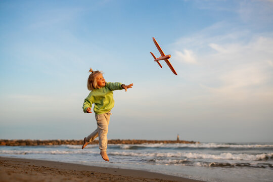 Smiling blond boy in green sweatshirt launching an orange toy airplane on the beach at sunset, looking at it as it flies above the sea.