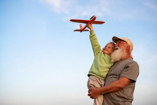 Joyful grandfather holding his happy blond grandson who is lifting a toy airplane into the sky, both enjoying playful time together under a clear blue sky.