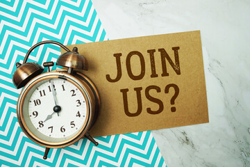 Join Us? text written on paper card with alarm clock on marble background