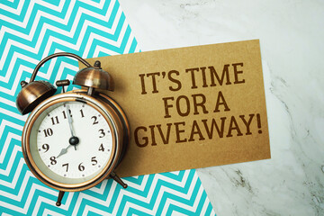 It's Time For A Giveaway! text written on paper card with alarm clock on marble background