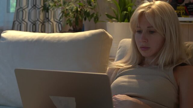 A young woman with blond hair sits on the sofa and looks at the laptop.