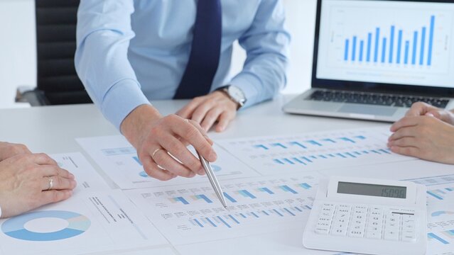 Business professional pointing at printed charts with a pen during a meeting, laptop showing bar graphs and calculator on table while team collaborates on financial analysis and planning - Powered by Adobe