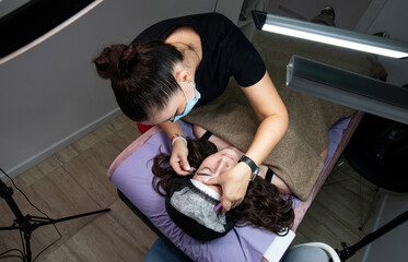A female specialist wearing a mask performs an eyelash extension procedure in a beauty salon