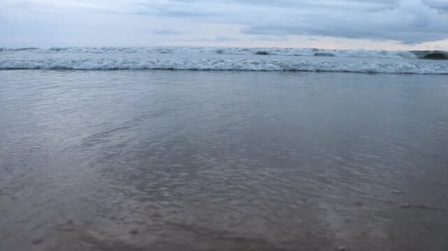 Ondas suaves tocam a areia molhada sob um c&eacute;u nublado azul-acinzentado, refletindo a calmaria do mar ao entardecer.