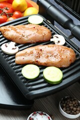Electric grill with raw marinated chicken fillet, mushrooms, spices and vegetables on wooden table, closeup