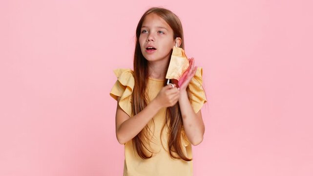 Young child girl fearfully shows a stop gesture while wearing a medical mask, then removes it and smiles with visible relief. School kid on pink background, portraying transition from fear to joy.