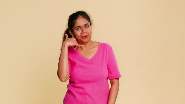Indian woman playfully forms call me gesture by placing hand near ear, smiling brightly. Hindu girl on beige background happily suggesting phone call, communication, casual chat, or friendly contact.