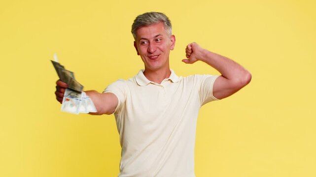 Middle-aged man in excitement holding bundle of dollars cash banknotes, celebrating lottery win casino jackpot or game success. Young guy expressing happiness and triumph isolated on yellow background