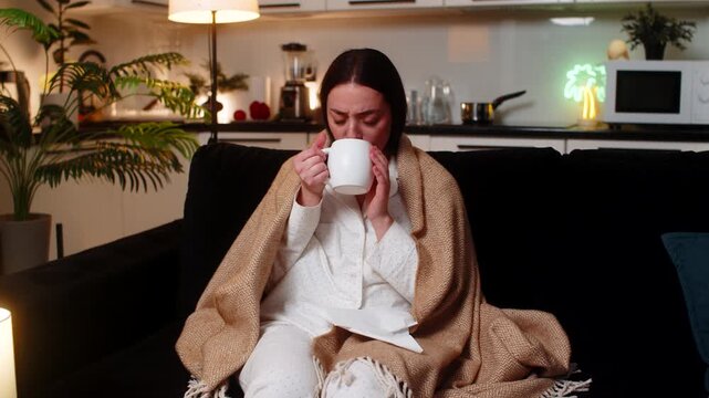Young woman at night home under blanket with hot tea and medicine sips for sore throat cough. Caucasian girl on sofa at evening uses tissue sneezes feels fever fatigue so rests quiet asks help softly.