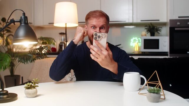 Young man at night home reads news on smartphone smiles celebrates online game jackpot win feels joy. Caucasian guy on sofa at table looks at phone raises fist happy reaction enjoys victory calm mood.