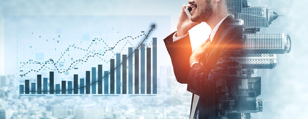 A businessman engages in a phone conversation while surrounded by financial graphs depicting...