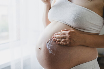 A pregnant woman applies skincare cream to her skin. This moment emphasizes self-care during pregnancy.