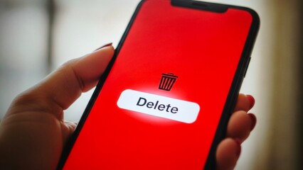 Closeup of a hand holding a smartphone with a red screen showing a delete button and trash can icon