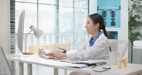 Asian female doctor in white coat sitting at desk in clinical office hospital, typing on computer,...