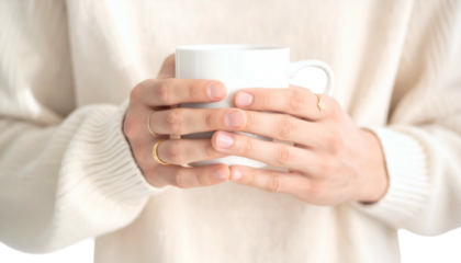 Close up of hands holding a white mug, wearing rings, cozy knit sweater in background