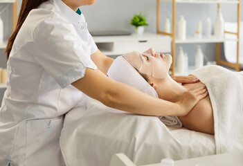 An esthetician gives a facial massage and works the shoulders as a sheet mask rests in a modern...