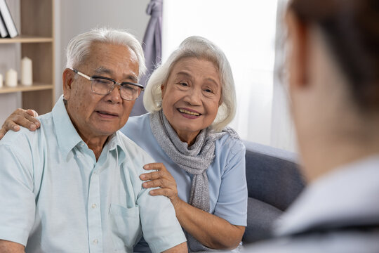 Asian elderly couple smiling during home medical consultation with caregiver present, woman touching partner shoulder showing affection and support