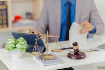 A lawyer or legal professional sits at a desk. On the desk are several objects related to law and a laptop, a clipboard with documents, a hammer, and a statue of the Goddess of Justice.