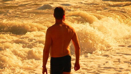 Man Ocean Sunset: Man watching golden waves crash at ocean beach during beautiful evening sunset for relaxation.