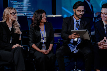 A large IT department meeting in a command center (SOC). An Indian analyst holds a tablet, listening with his team to a cybersecurity update on an AI system.