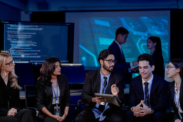 Obraz premium An Indian man with a tablet asks a question during a large team briefing. His diverse colleagues, including Indian and Caucasian women and men, listen in a high-tech conference room.