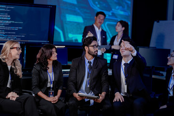A large IT department meeting in a command center (SOC). An Indian analyst holds a tablet,...