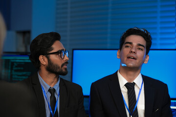 A diverse team, an Indian man with glasses and a Caucasian man, listens intently during a professional business meeting or conference in a modern, high-tech office at night.